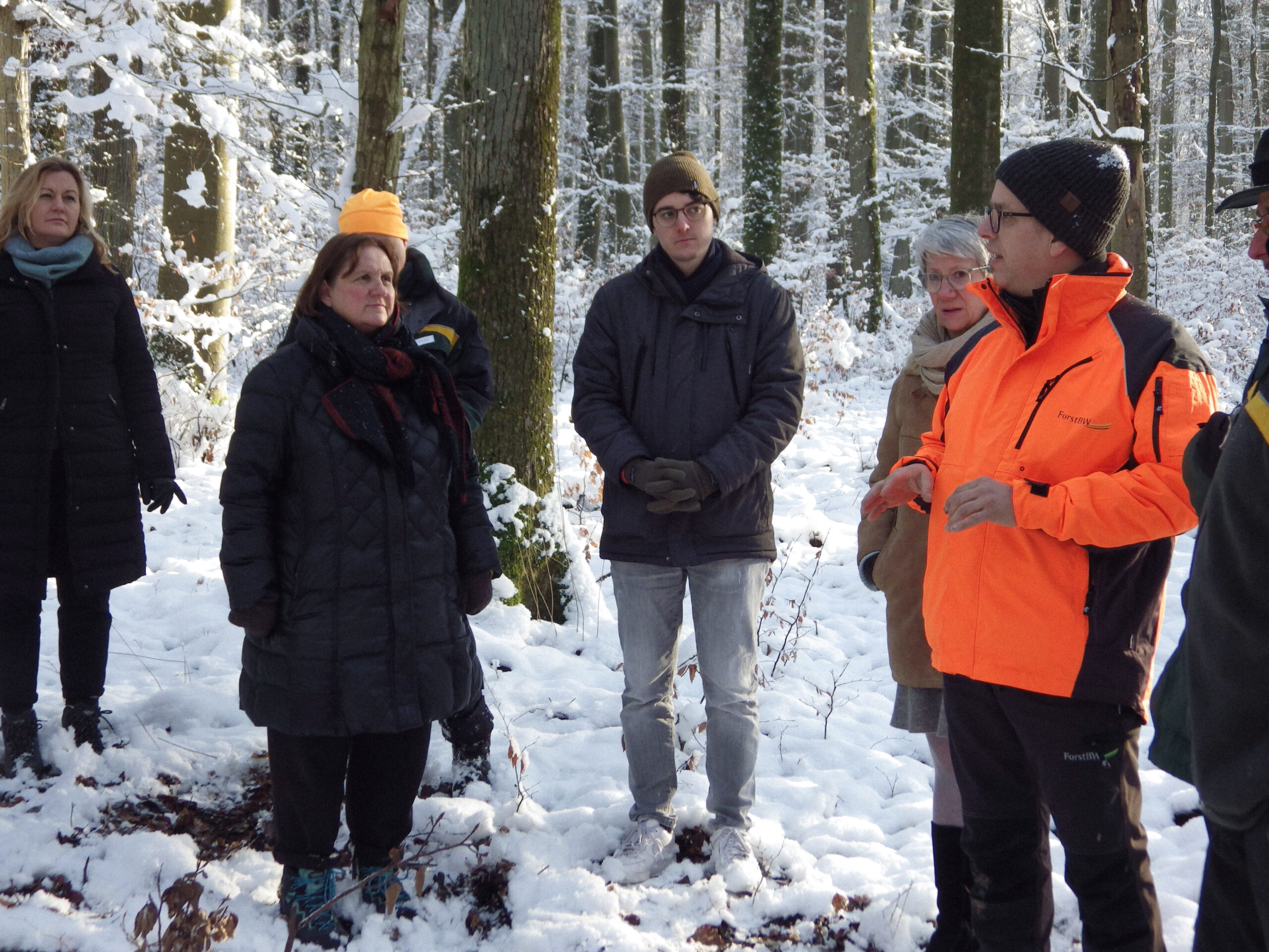 Theresa Schopper, Mario Dietel und Catherine Kern hören einem Förster zu. Sie stehen in einem verschneiten Wald.