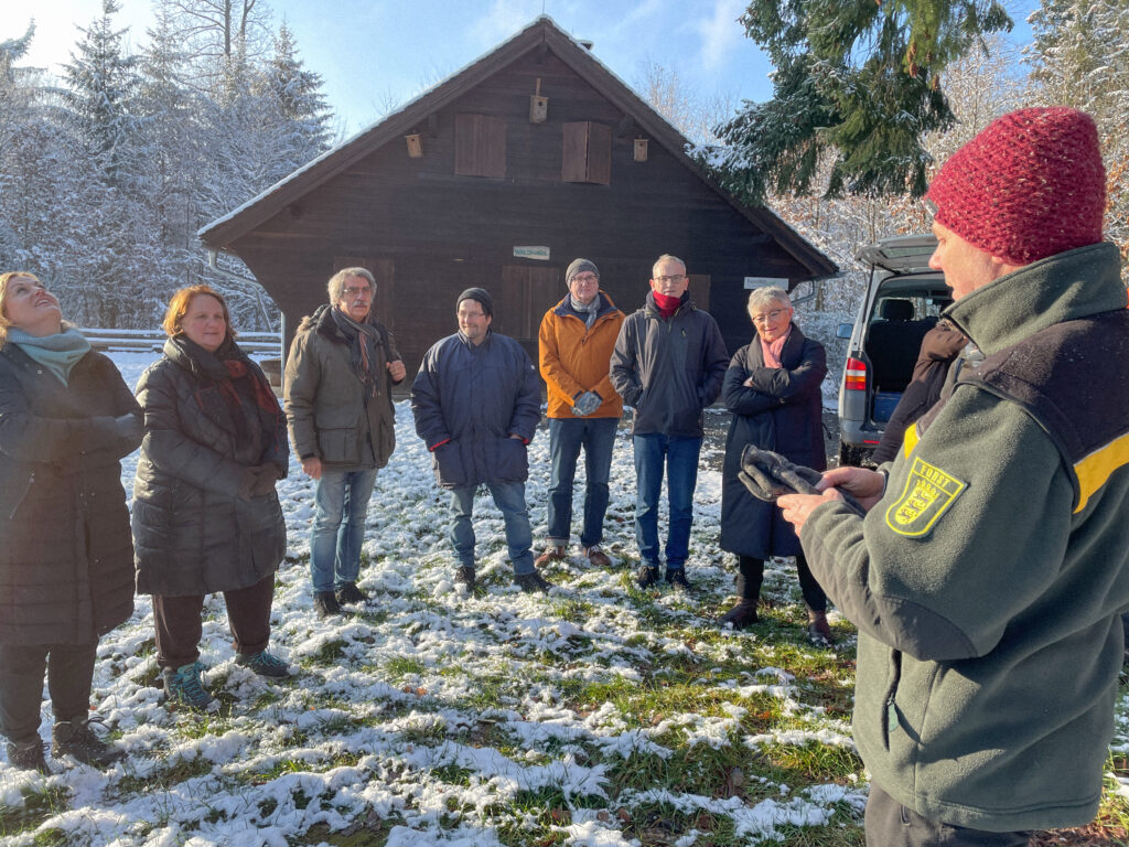Uli Baumhäuser erklärt der Delegation um Theresa Schopper im Wald vor einer Hütte, wie er Waldpädagogik versteht